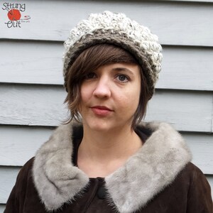 May include: A handmade, textured crochet hat in shades of cream, gray, and brown. The hat has a ribbed brim and a rounded crown. The image also shows a brown coat with a gray fur collar. The logo "Strung Out Fiber Arts" is in the top left corner.