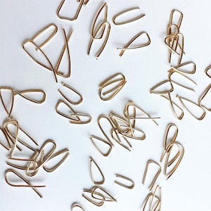 May include: A collection of small, gold-colored metal hair pins scattered on a white surface. The pins have various shapes, including U-shapes, teardrops, and looped designs. The pins are made of a shiny, metallic material.