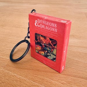 Op de afbeelding: Een rode miniatuurdoos-sleutelhanger met het "Dungeons & Dragons"-logo en een drakenillustratie. De sleutelhanger heeft een zwarte metalen ring en ketting. Op de doos staat "SET 1 BASIC RULES".