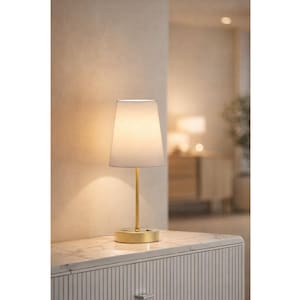 May include: A small table lamp with a white lampshade and a gold-colored base and stem. The lamp is turned on, emitting a warm glow. The lamp sits on a white marble-topped cabinet.