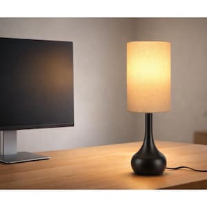 May include: A black table lamp with a beige cylindrical lampshade. The lamp has a curved base and sits on a wooden desk. A large black computer monitor is visible in the background.
