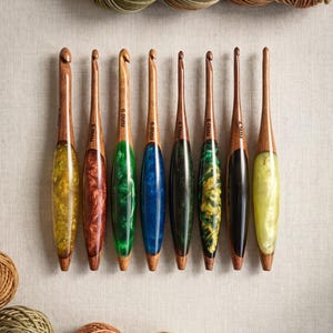 May include: A collection of eight handcrafted crochet hooks with wooden handles and colorful resin accents. The hooks are arranged in a row, showcasing a variety of colors including yellow, red, green, blue, and black. Yarn skeins and balls of yarn are also visible.