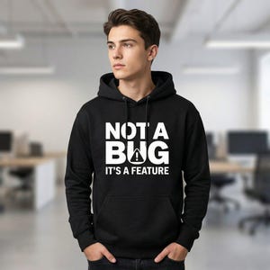 Op de afbeelding: Zwarte hoodie met de witte tekst "NOT A BUG IT'S A FEATURE". De hoodie heeft een capuchon met trekkoord en een voorzak. Het ontwerp bevat een uitroepteken in een waarschuwingsdriehoek.