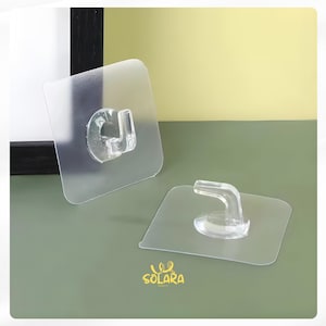 May include: Two clear, adhesive wall hooks with a square base and a curved hook. The hooks are made of transparent plastic for easy installation. The logo "SOLARA PARTY" is visible in the bottom right corner.