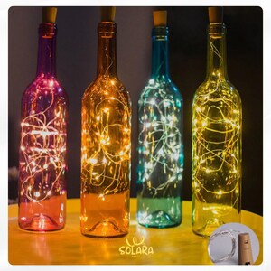 May include: Four decorative wine bottles in purple, orange, blue, and yellow, each filled with string lights. The bottles have cork stoppers and are arranged on a yellow surface. The word "SOLARA" is visible.