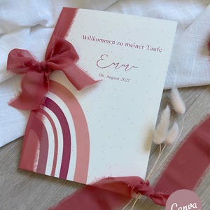 Puede incluir: Una tarjeta de invitación de bautizo con un diseño de arcoíris en acuarela en tonos rosas y rojos. La tarjeta presenta el texto "Willkommen zu meiner Taufe" y el nombre "Emma" con la fecha "06. August 2027". Una cinta y un lazo a juego adornan la tarjeta.