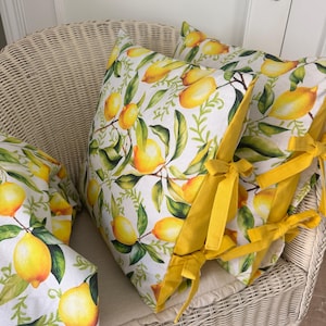 Puede incluir: Cojines blancos con un estampado de limones y hojas. Los cojines tienen lazos amarillos y están colocados en una silla de mimbre blanca. El diseño de limón presenta limones amarillos y hojas verdes.