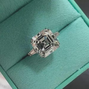 May include: A square-cut diamond ring with a silver band, presented in a teal ring box. The diamond features a multi-faceted design, reflecting light. The ring is a piece of jewellery.