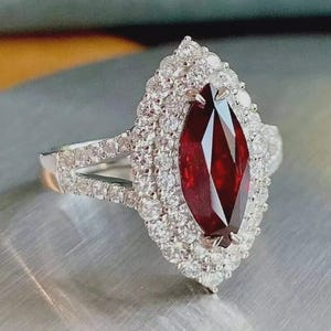 May include: A marquise-cut, deep red gemstone ring with a double halo of small, clear gemstones. The ring's band is silver-colored, and the setting is intricate, showcasing the central stone.