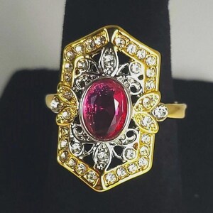 May include: A gold-toned ring with a large oval ruby center stone. The ring features a decorative filigree design with clear accent stones and a geometric frame. The ring is set against a black background.