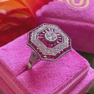 Könnte beinhalten: Ein Ring im Art-Deco-Stil mit geometrischem Design. Der Ring hat einen zentralen runden Diamanten, der von kleineren Rubinen und Diamanten umgeben ist, gefasst in einem Band aus Silber oder Weißgold. Der Ring wird auf einer rosa Samtoberfläche präsentiert.