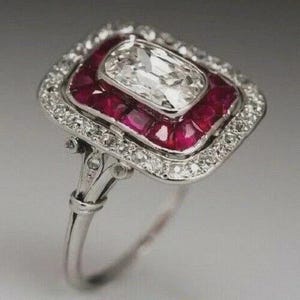Puede incluir: Un anillo de estilo vintage con una gran piedra preciosa rectangular y transparente en el centro, rodeada por un halo de diamantes más pequeños y brillantes. El anillo presenta un borde de piedras preciosas rojo vibrante y una banda plateada.
