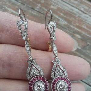 May include: Elegant drop earrings featuring a central round diamond surrounded by a halo of red gemstones. The earrings have a silver-tone metal setting with intricate detailing and additional small diamonds. The design includes a floral-inspired top and a hook closure.