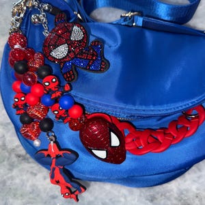 May include: A blue satin crossbody bag adorned with Spider-Man themed charms. The bag features a red chain strap and multiple Spider-Man character charms in red, black, and blue. The bag has a gold zipper and a blue strap.