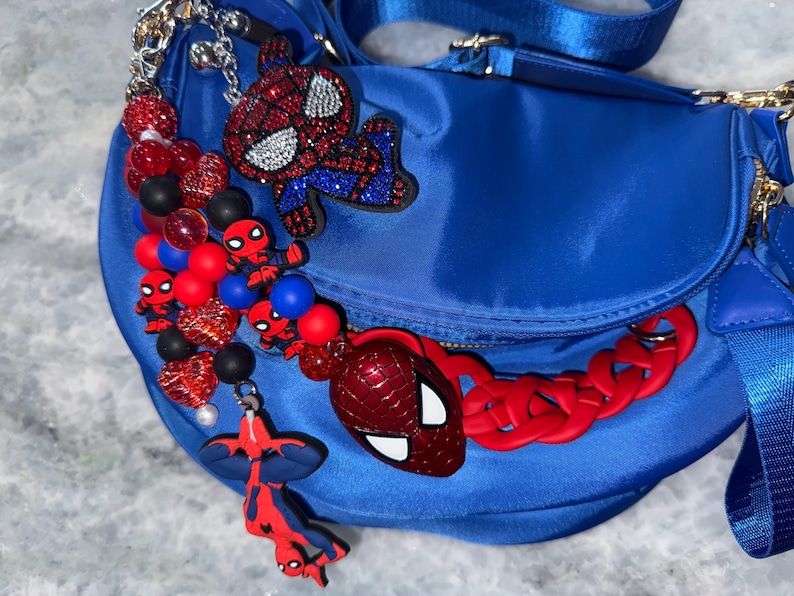 May include: A blue satin crossbody bag adorned with Spider-Man themed charms. The bag features a red chain strap and multiple Spider-Man character charms in red, black, and blue. The bag has a gold zipper and a blue strap.