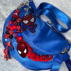 May include: A royal blue fanny pack with a matching adjustable strap. The bag is adorned with multiple Spider-Man themed charms, including beaded accents in red, black, and blue, and a red chain.