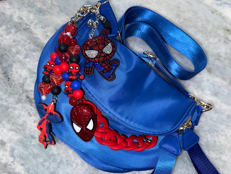 May include: A royal blue fanny pack with a matching adjustable strap. The bag is adorned with multiple Spider-Man themed charms, including beaded accents in red, black, and blue, and a red chain.
