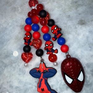 May include: A beaded keychain featuring a Spider-Man theme. The keychain includes red, blue, and black beads, along with Spider-Man character charms and a Spider-Man mask pendant. The design incorporates a mix of textures and shapes.