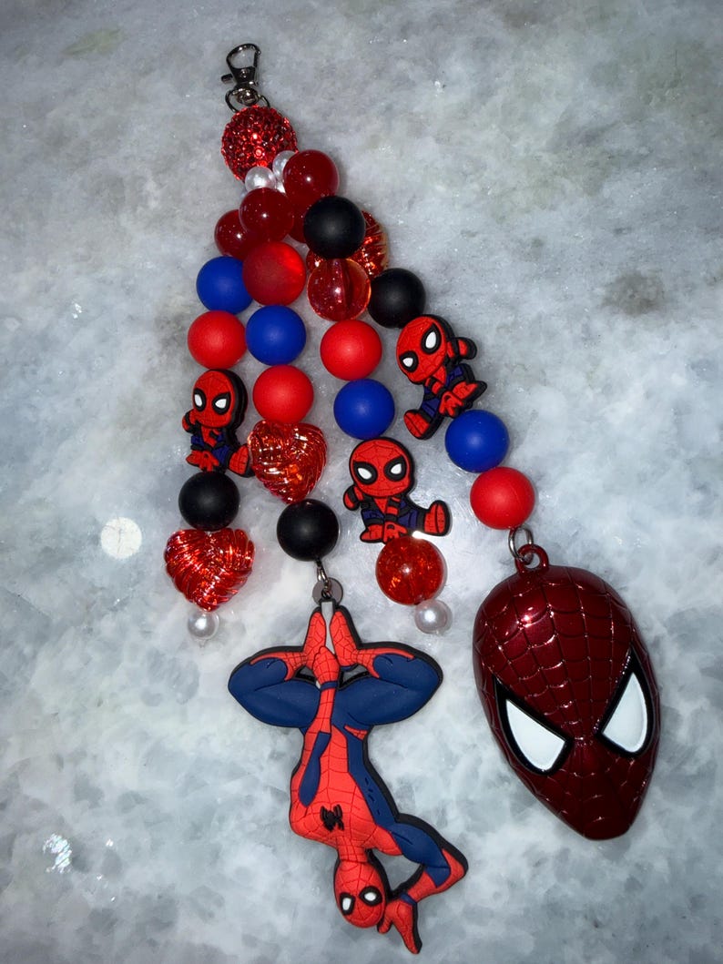 May include: A beaded keychain featuring a Spider-Man theme. The keychain includes red, blue, and black beads, along with Spider-Man character charms and a Spider-Man mask pendant. The design incorporates a mix of textures and shapes.