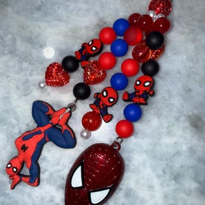 May include: A Spider-Man themed keychain featuring a red and blue Spider-Man figure, a red Spider-Man mask, and various red, blue, and black beads. The keychain includes a silver clasp for easy attachment.