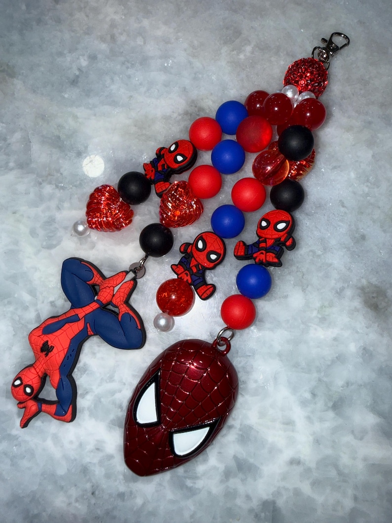 May include: A Spider-Man themed keychain featuring a red and blue Spider-Man figure, a red Spider-Man mask, and various red, blue, and black beads. The keychain includes a silver clasp for easy attachment.
