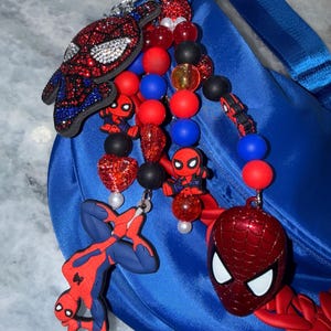 May include: A blue bag with a strap is adorned with a collection of Spider-Man themed charms. The charms include a bedazzled Spider-Man mask, various Spider-Man figures, and beaded accents in red, blue, black, and clear.