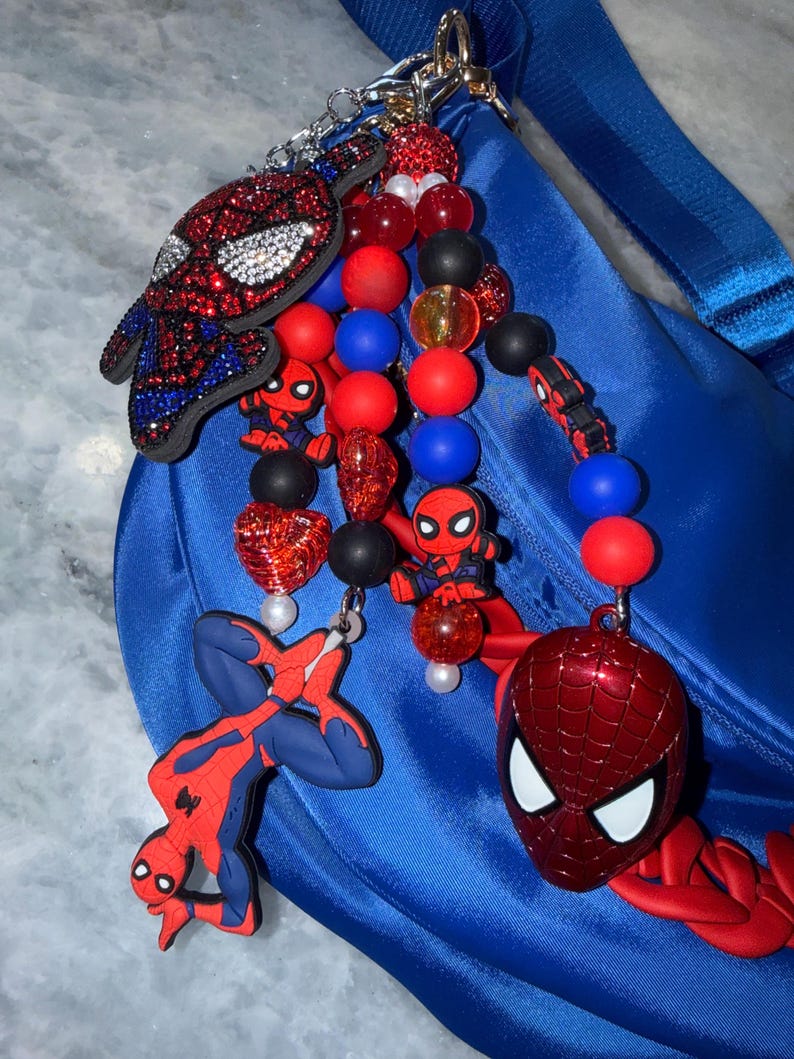 May include: A blue bag with a strap is adorned with a collection of Spider-Man themed charms. The charms include a bedazzled Spider-Man mask, various Spider-Man figures, and beaded accents in red, blue, black, and clear.