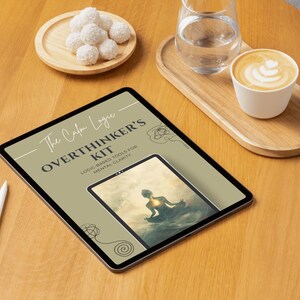 Może przedstawiać: Tablet cyfrowy wyświetlający "The Calm Logic OVERTHINKER'S KIT" z mniejszym obrazem osoby medytującej. Tablet leży na drewnianej powierzchni, w towarzystwie filiżanki kawy, szklanki wody i talerza z białymi, okrągłymi smakołykami.