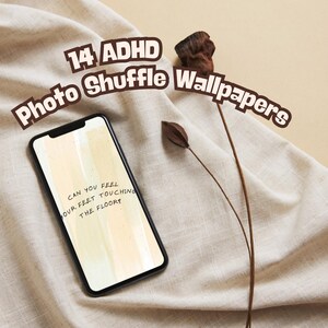May include: A smartphone with a black frame displays a light yellow and white abstract design with the text "CAN YOU FEEL OUR FEET TOUCHING THE FLOOR?". The image is set against a beige fabric background with the text "14 ADHD Photo Shuffle Wallpapers".