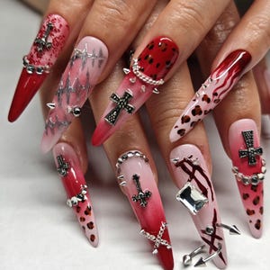 May include: Long, pointed acrylic nails with a red to pink gradient. The nails are decorated with silver studs, crosses, chains, and a leopard print pattern. Some nails have a silver barbed wire design.