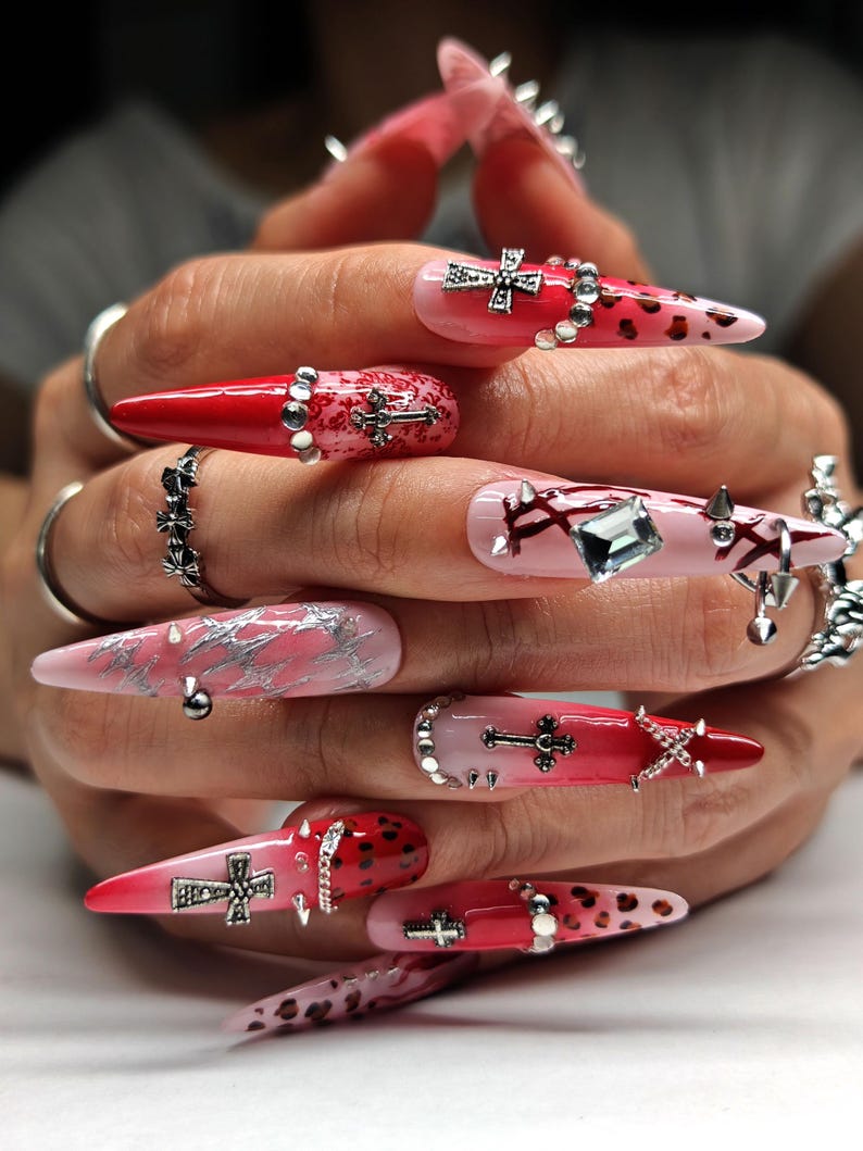 May include: Long, pointed fingernails with a gradient of red and pink polish. The nails are decorated with silver crosses, rhinestones, and studs. Some nails have a leopard print design. Silver rings are worn on the fingers.