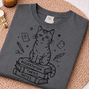 May include: Dark gray t-shirt featuring a grumpy cat illustration sitting on a stack of books labeled "Dissertation", "Thesis", and "Notes". The design includes a pencil, stars, and floating papers.