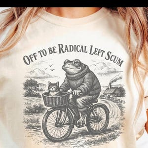 May include: Beige t-shirt with a graphic of a frog wearing a sweater and glasses riding a bicycle with a cat in a basket. The text "OFF TO BE RADICAL LEFT SCUM" is above the frog.