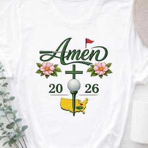 Peut inclure: T-shirt blanc avec un motif sur le thème du golf. Le motif comprend le mot "Amen" en script vert, une balle de golf sur un tee, une croix, des fleurs roses, les chiffres "20" et "26" et une carte jaune des États-Unis avec un drapeau de golf.