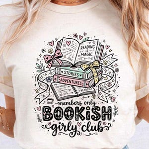 Peut inclure: T-shirt crème avec un motif fantaisiste de livres empilés, des cœurs et des nœuds. Le texte comprend "Reading is Magic", "Stories", "Adventures" et "Bookish Girly Club" dans une police décorative.
