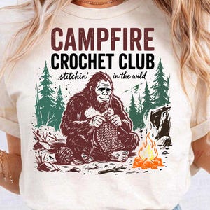 May include: Cream-colored t-shirt with a graphic featuring a Bigfoot crocheting by a campfire. The text reads "Campfire Crochet Club" with "stitchin' in the wild" below. The design includes trees, a campfire, and a ball of yarn.