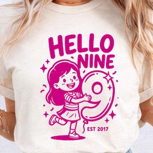 Peut inclure: T-shirt crème avec un motif magenta. Le motif présente le texte "HELLO NINE" au-dessus d'une fillette de dessin animé tenant un ballon en forme de chiffre neuf. Le texte "EST 2017" est en dessous.