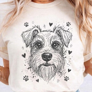 Puede incluir: Camiseta color crema con un boceto detallado en blanco y negro de la cara de un perro. El diseño incluye corazones, huellas de patas y estrellas. La camiseta tiene mangas enrolladas.