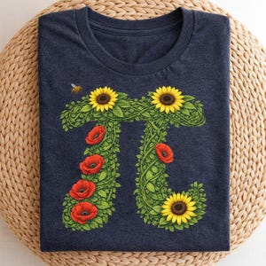 Può includere: T-shirt blu navy con un design floreale del simbolo Pi. Il simbolo Pi è formato da viti verdi, girasoli gialli e papaveri rossi. Una piccola ape vola vicino alla parte superiore della maglietta. La maglietta è piegata su una superficie tessuta.