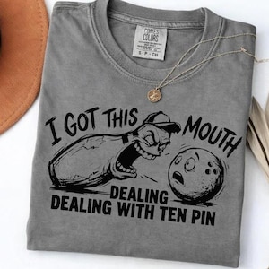 Peut inclure: T-shirt gris avec un motif sur le thème du bowling. Le motif représente une quille de bowling de dessin animé avec un visage en colère et une boule de bowling avec une expression surprise. Le texte dit "I GOT THIS MOUTH DEALING WITH TEN PIN."