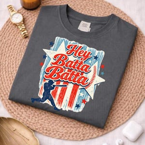 May include: A gray t-shirt featuring the phrase "Hey Batta Batta" in red and white script, set against a star-shaped design with red and blue stripes and stars. A baseball player silhouette is also featured.