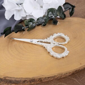 May include: Silver-colored decorative scissors with ornate handles, resting on a wooden surface. White flowers and greenery are arranged nearby. The scissors have a detailed, vintage design, suitable for crafting or decorative purposes.