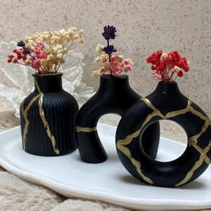 May include: Three matte black vases with gold-coloured crack detailing, each holding small dried flowers in shades of pink, white, and purple. The vases include a ribbed cylindrical shape, a curved arch form, and a circular donut-like design.