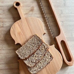 Könnte beinhalten: Ein Brotschneidebrett und Brotsäge-Set aus Holz. Das Schneidebrett hat die Form einer Geige, mit drei Brotscheiben darauf. Die Brotsäge hat einen Holzgriff und eine gezahnte Klinge. Das Set ist aus hellbraunem Holz gefertigt.
