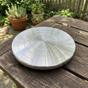 Puede incluir: Una rueda de alfarero redonda y plateada con círculos concéntricos. La rueda es de metal y está sobre una mesa de madera envejecida. El fondo incluye plantas en macetas y una valla de madera.