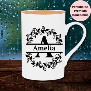 May include: A white bone china mug featuring a black graphic design of a circular floral wreath surrounding a large letter A. The name Amelia is printed in black text across the centre of the letter. A pink circular badge reads Personalise Premium Bone China.