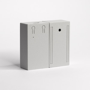 Könnte beinhalten: Ein hellgraues, rechteckiges Objekt mit strukturierter Oberfläche. Das Objekt besteht aus zwei verbundenen Abschnitten. Ein Abschnitt hat zwei schlüssellochförmige Aussparungen und einen kleinen Punkt. Der andere Abschnitt hat ein kreisförmiges Loch und zwei vertikale Linien.