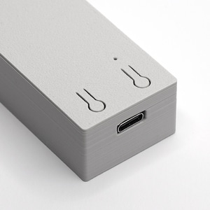 Könnte beinhalten: Nahaufnahme eines rechteckigen, hellgrauen elektronischen Geräts. Das Gerät hat einen USB-C-Anschluss und zwei schlüssellochförmige Aussparungen auf der Oberseite. Das Gerät besteht aus einem strukturierten Material.