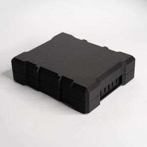 Könnte beinhalten: Eine schwarze, rechteckige Box mit strukturierter Oberfläche. Die Box hat eine Reihe vertikaler Linien auf einer Seite und ein geometrisches Design. Die Box steht auf einer weißen Oberfläche.