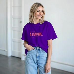 Könnte beinhalten: Ein lila T-Shirt mit dem Text "NOT-A-BUG A FEATURE. working as intended" in Rosa. Das T-Shirt wird mit einer blauen Jeans getragen.